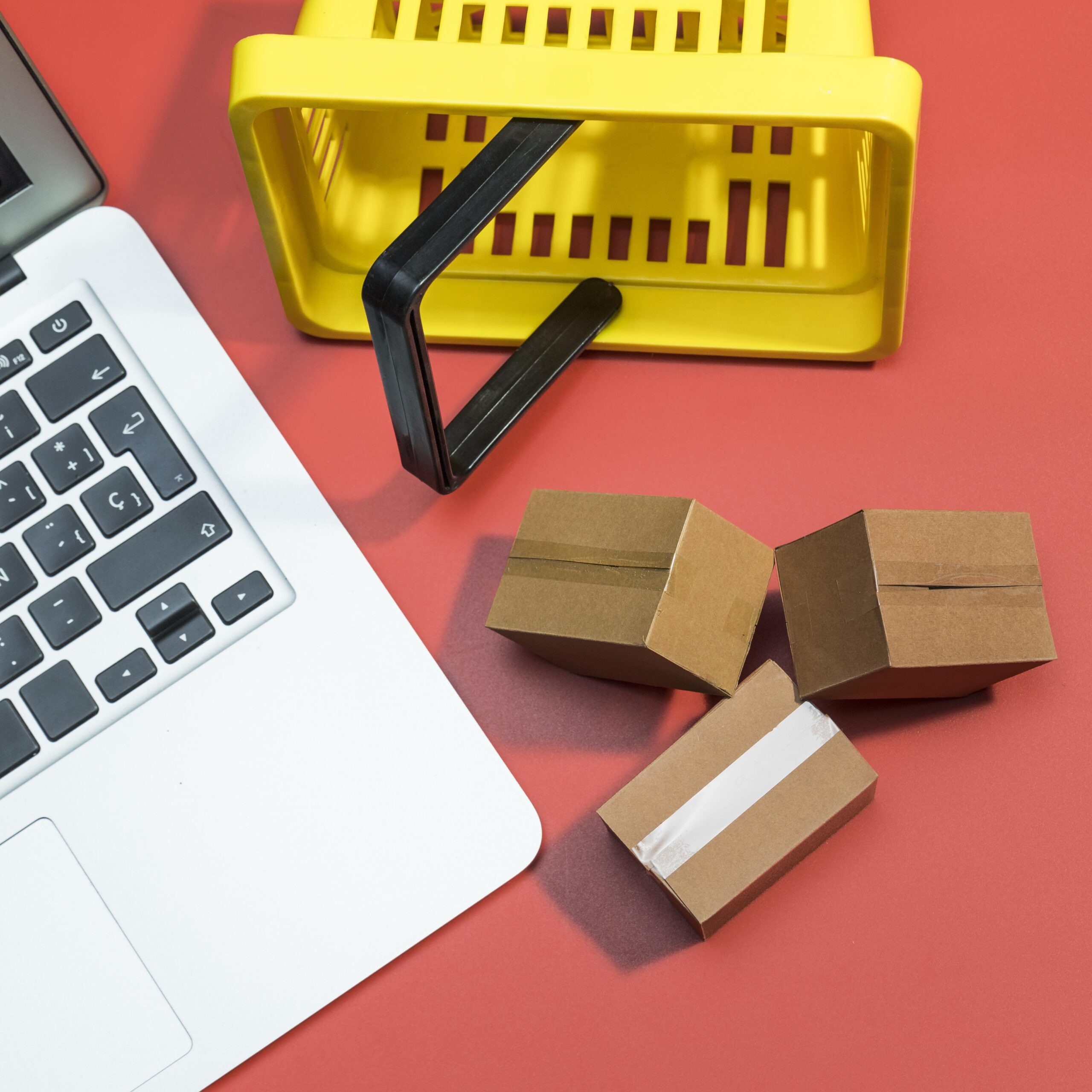 A laptop, miniature cardboard boxes, and a small shopping basket on a red surface, symbolizing efficient e-commerce logistics, supply chain optimization, and streamlined global trade solutions.