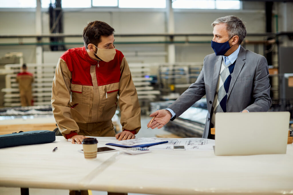 Two professionals wearing masks engaged in a discussion at a manufacturing facility, representing expert procurement, supply chain management, and tailored import-export solutions for businesses.